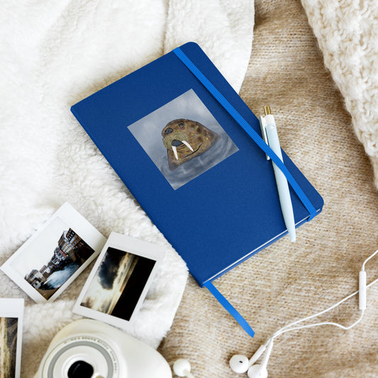 Walrus  on blue hardcover notebook/journal. Gentle giant, spirit guide, medicine man. Close up of Walrus head popping out of the sea. Product mockup shown laying on a bed with a pen tucked into the elastic enclosure, photos and disc player and earphones,