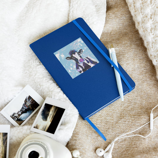 Cow Spirit, Divine Mother, graces the cover of a hardcover notebook, for morning pages, journaling, and note-taking. Shown with elastic closure holding a pen, on a bed with photos and electronics