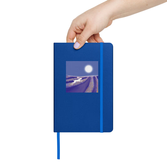 Hand holding a blue notebook with a hare running through lavender fields cover design