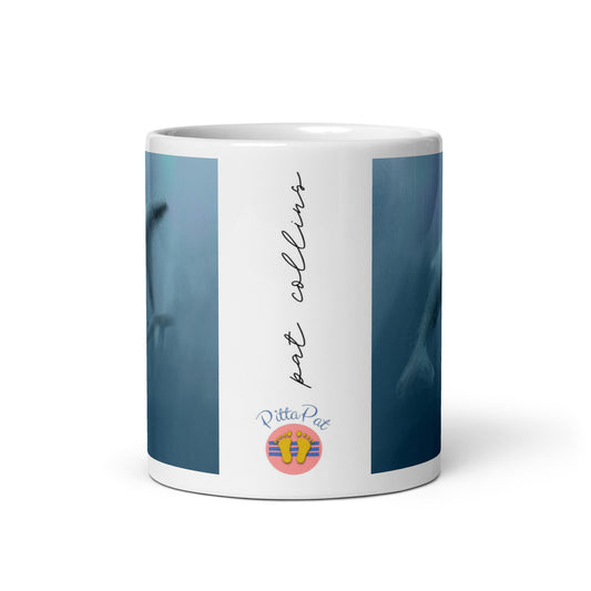 Side view of white ceramic mug with misty blue whale design. Side shows PittaPat logo and artist signature