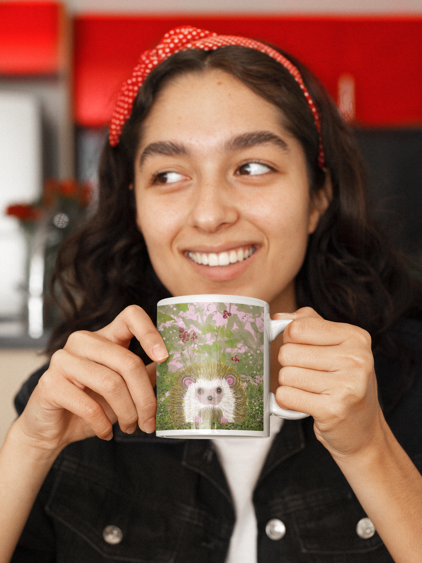 A woman sitting in a red kitchen and wearing a red bow in her hair is holding a mug with the design of a hedgehog in berry bushes.