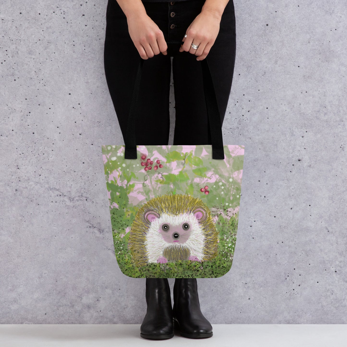 Woman holding a sturdy, washable tote bag with an image of the garden animal, the hedgehog.  Image shows hedgehog resting in a bramble of vines and berries and with a white butterfly. Hedgehog, with his sweet face, is gentle and kind spirit animal / spirit guide connected to Mother Earth. 15x15 inch. Black handles.