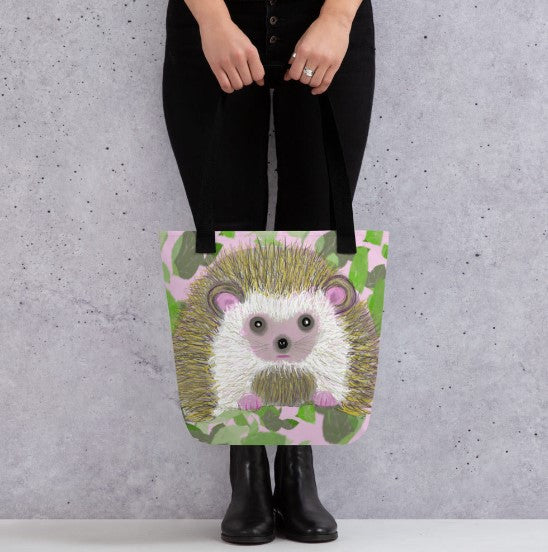 A hedgehog sitting in a field of pink with green leaves on this study tote bag. A tote bag to carry everything you need from office to the beach.