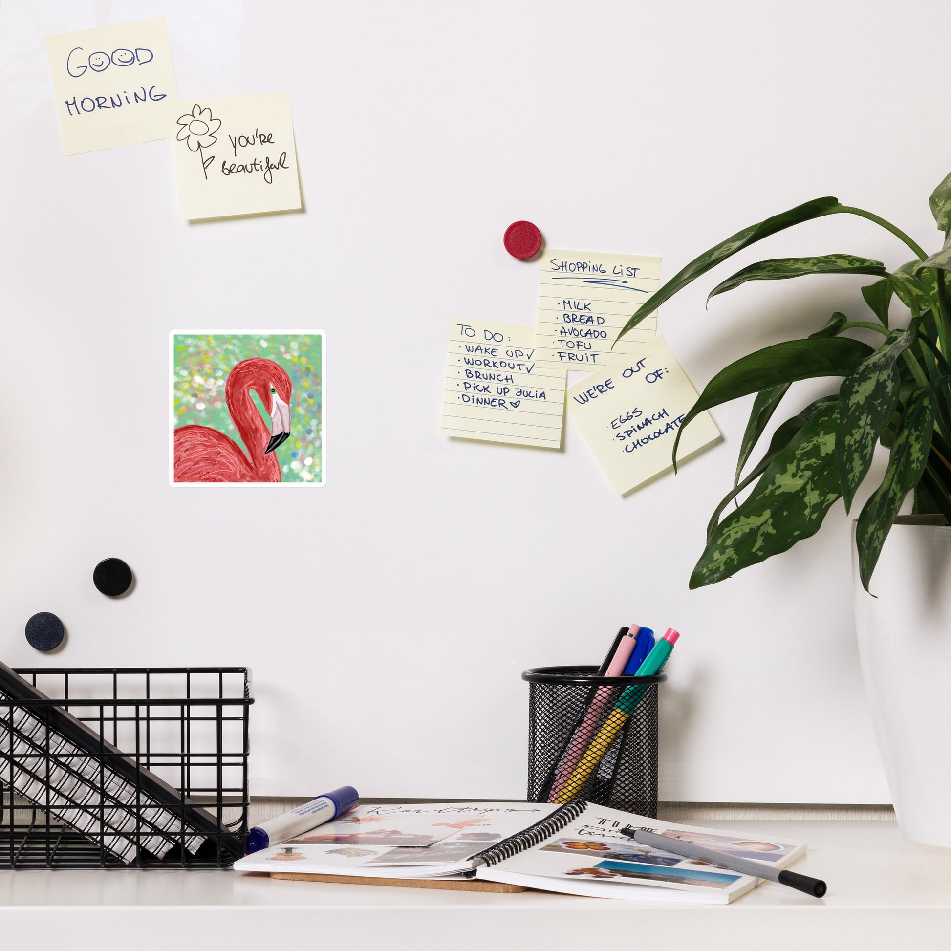 Pink Flamingo design on magnet on a magnetic board. Desk with stationery items, notes, and a plant on a white wall.