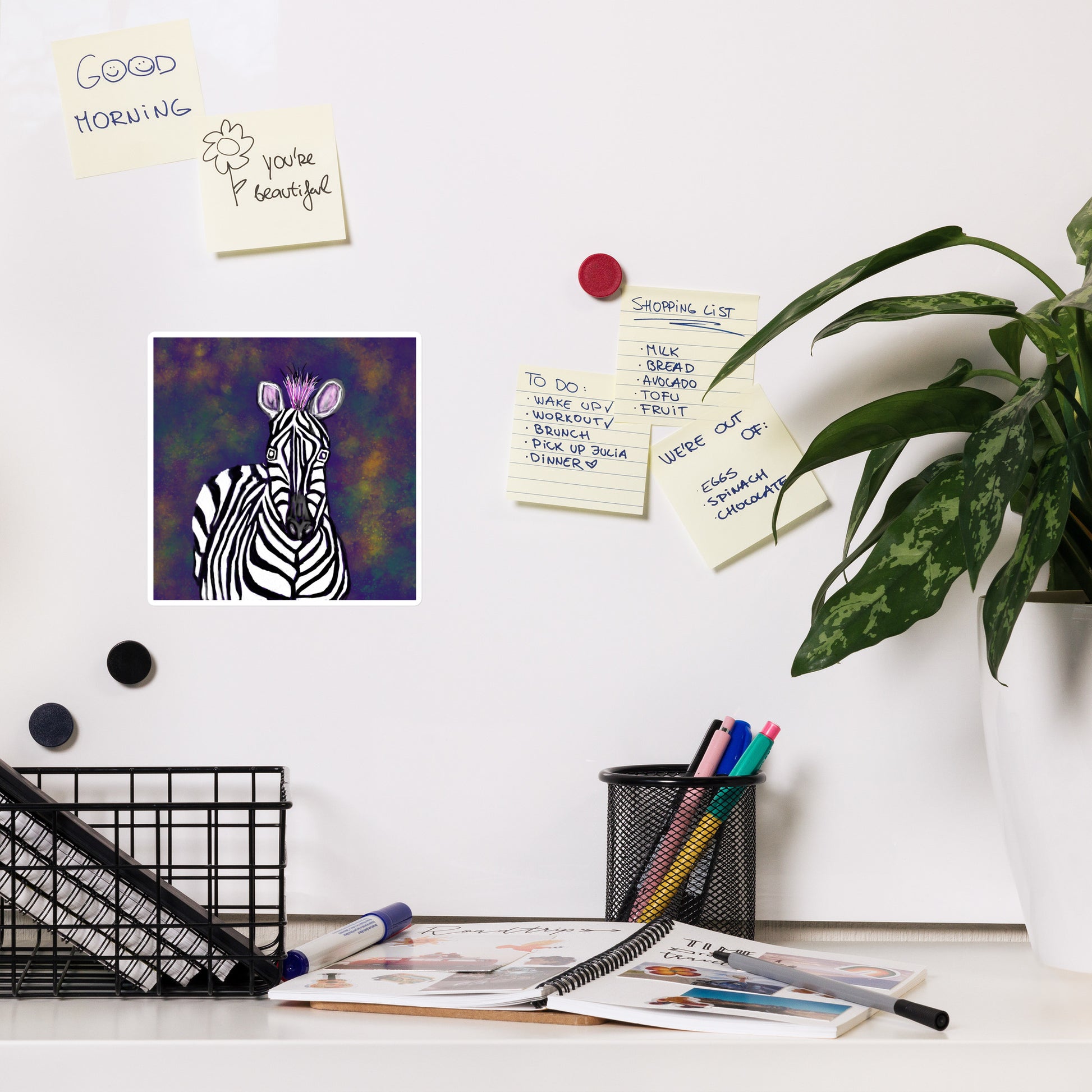 Desk with notes, pens, and stationery/ Above the desk is a zebra magnet on a white board .