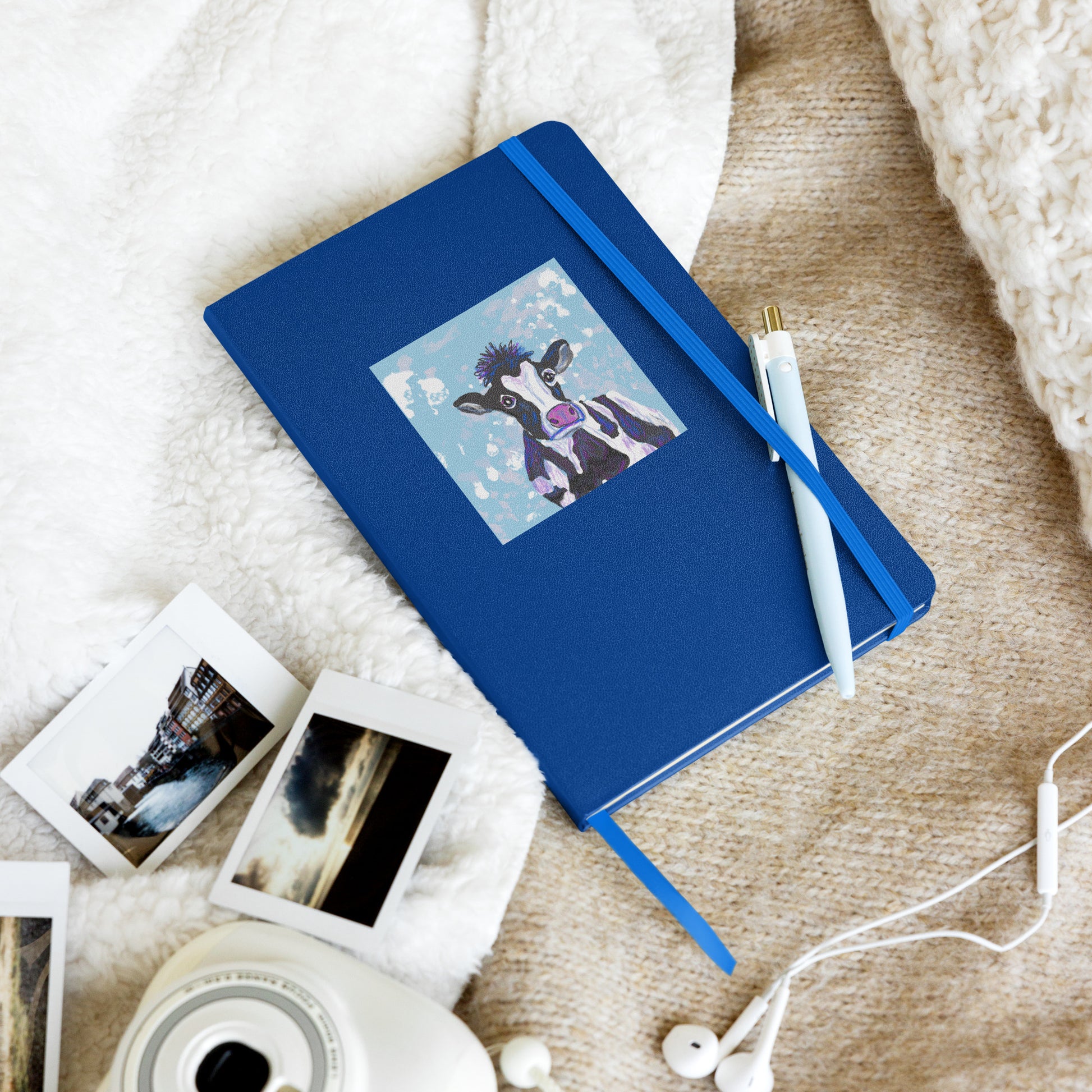 Cow Spirit, Divine Mother, graces the cover of a hardcover notebook, for morning pages, journaling, and note-taking. Shown with elastic closure holding a pen, on a bed with photos and electronics