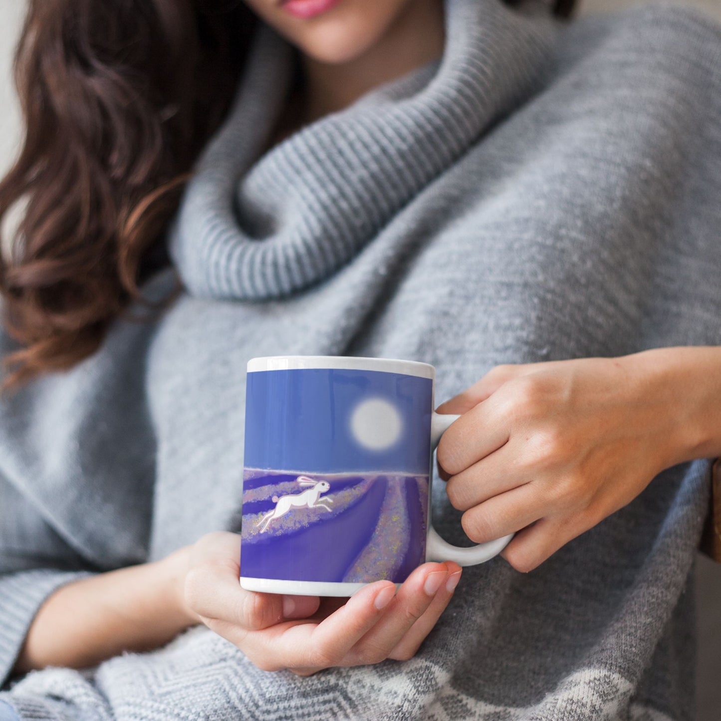 A woman in a gray sweater holding a wild hare spirit mug with an original  artists  design of a hare running through a lavender field under a full moon.