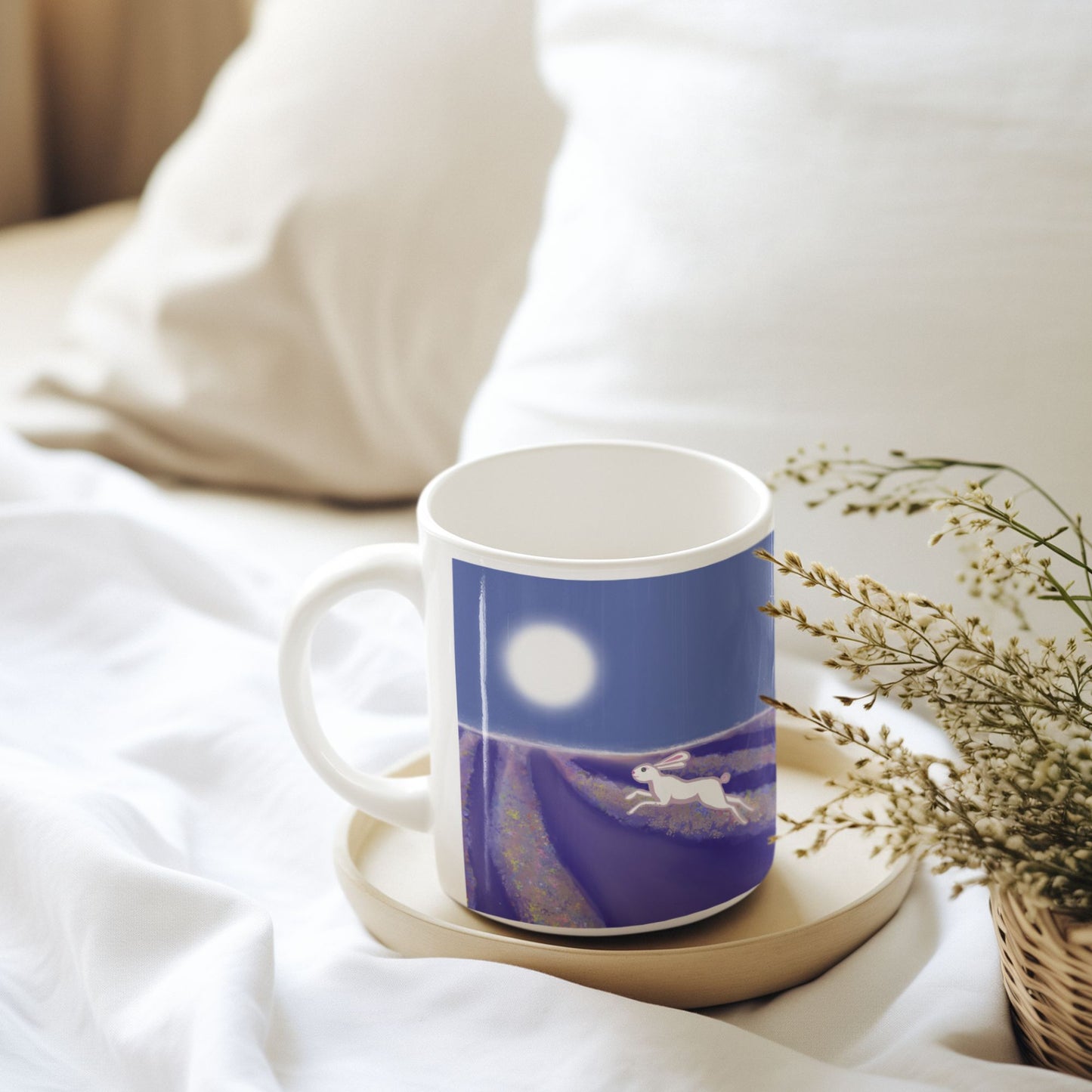 A white ceramic hare spirit mug with a design of a wild hare running through a lavender field under a full moon sits next to a basket of dried flowers on a bed with white pillows and comforter.