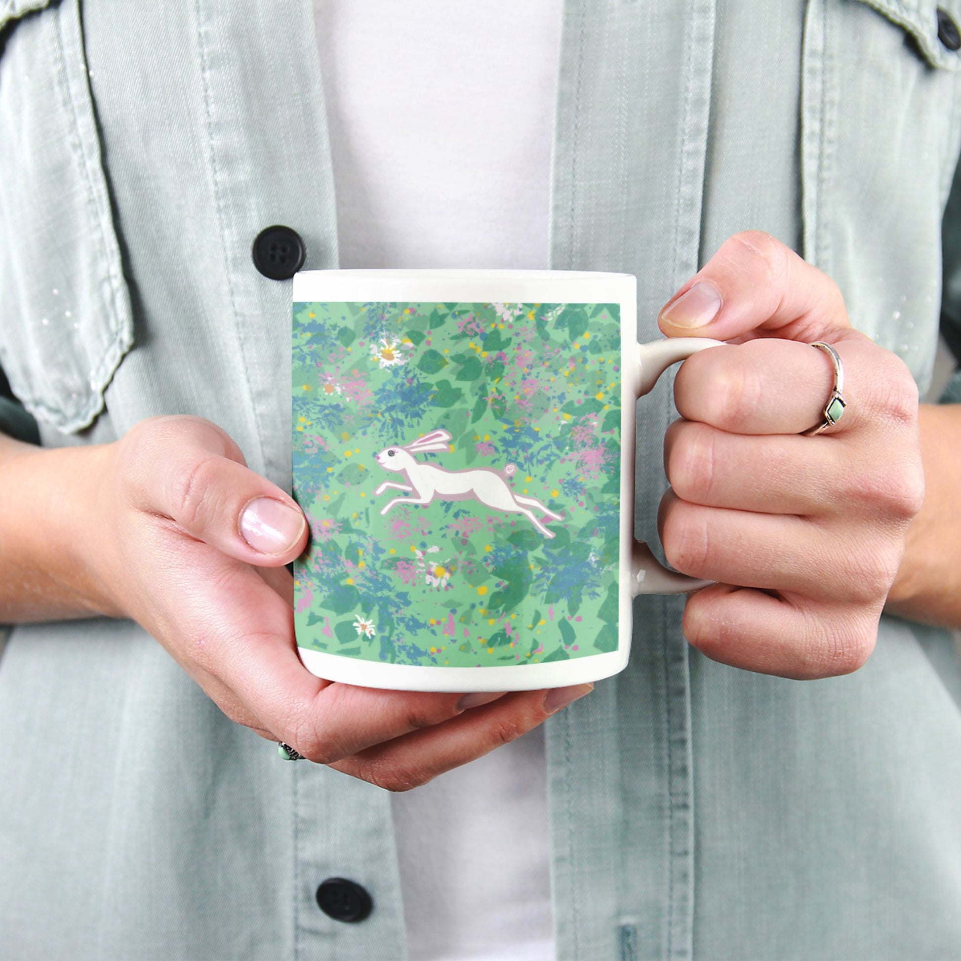 Person holding a mug with an image of a wild hare running through a field of flowers. 
