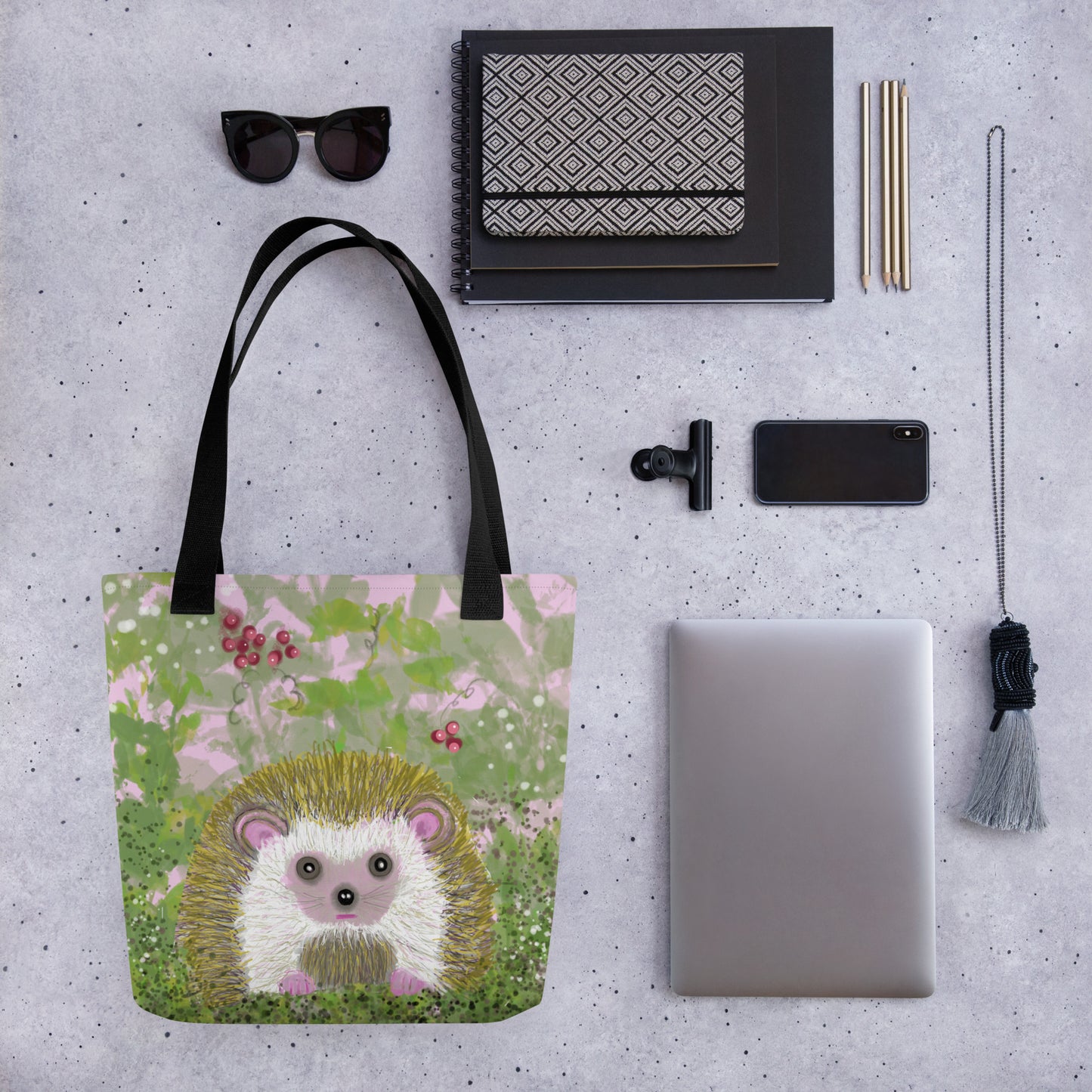 Tote bag with hedgehog design on a gray surface with various items often carried in a tote bag, laptop, notebook, phone, sun glasses, etc.