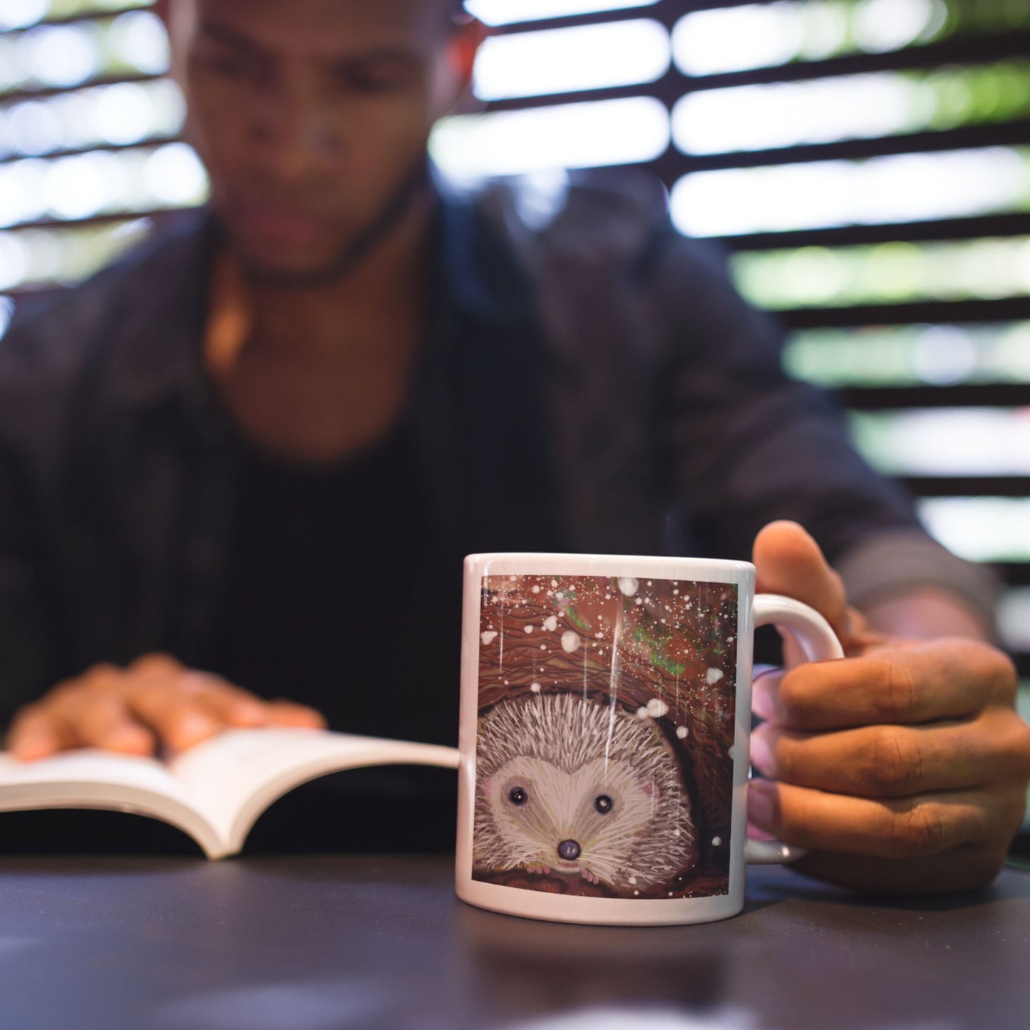 Person sitting at a table, reading a book with a mug featuring a hedgehog design.