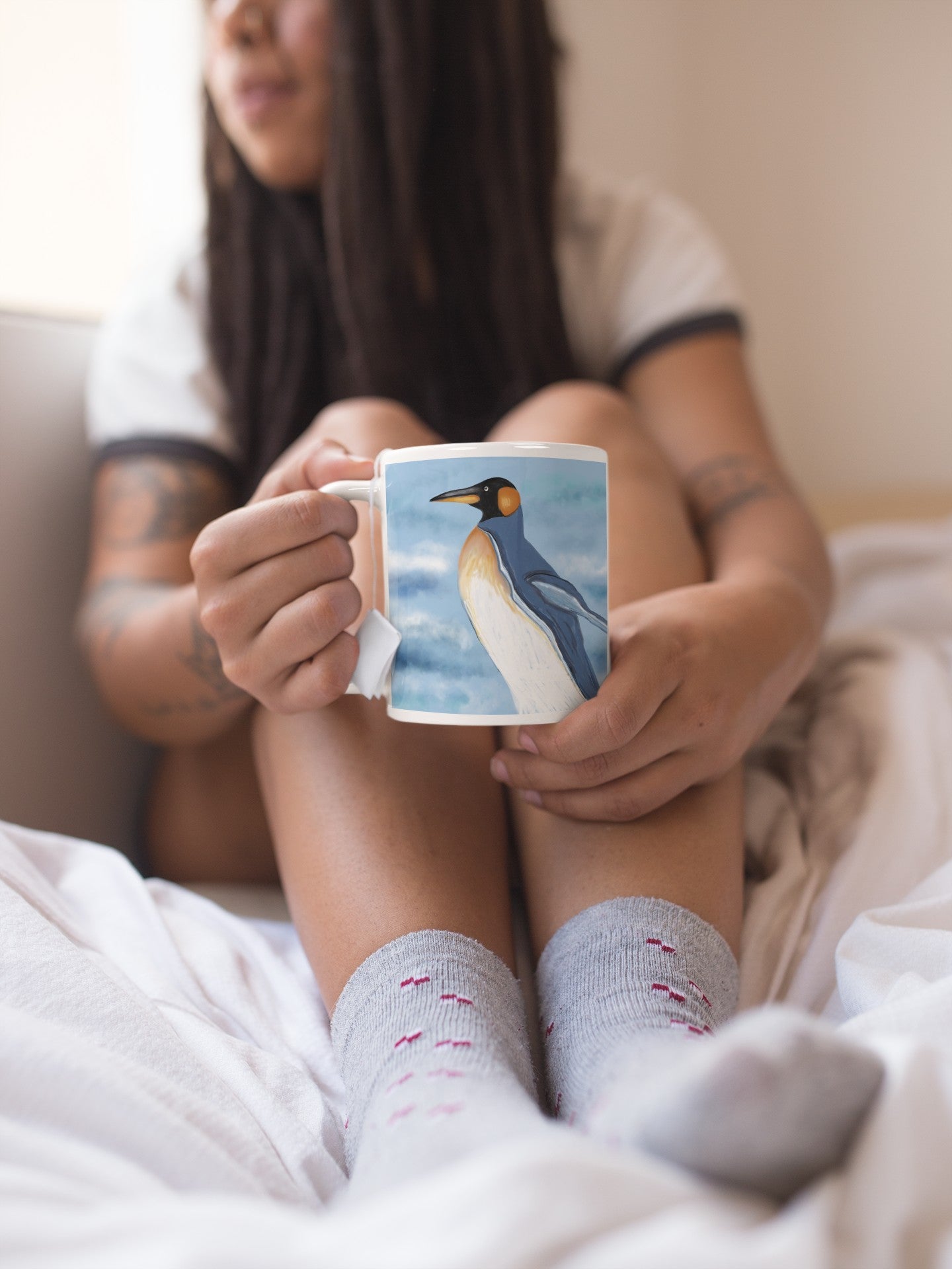 Woman holding a Penguin mug, sitting on a bed and staring out a window.