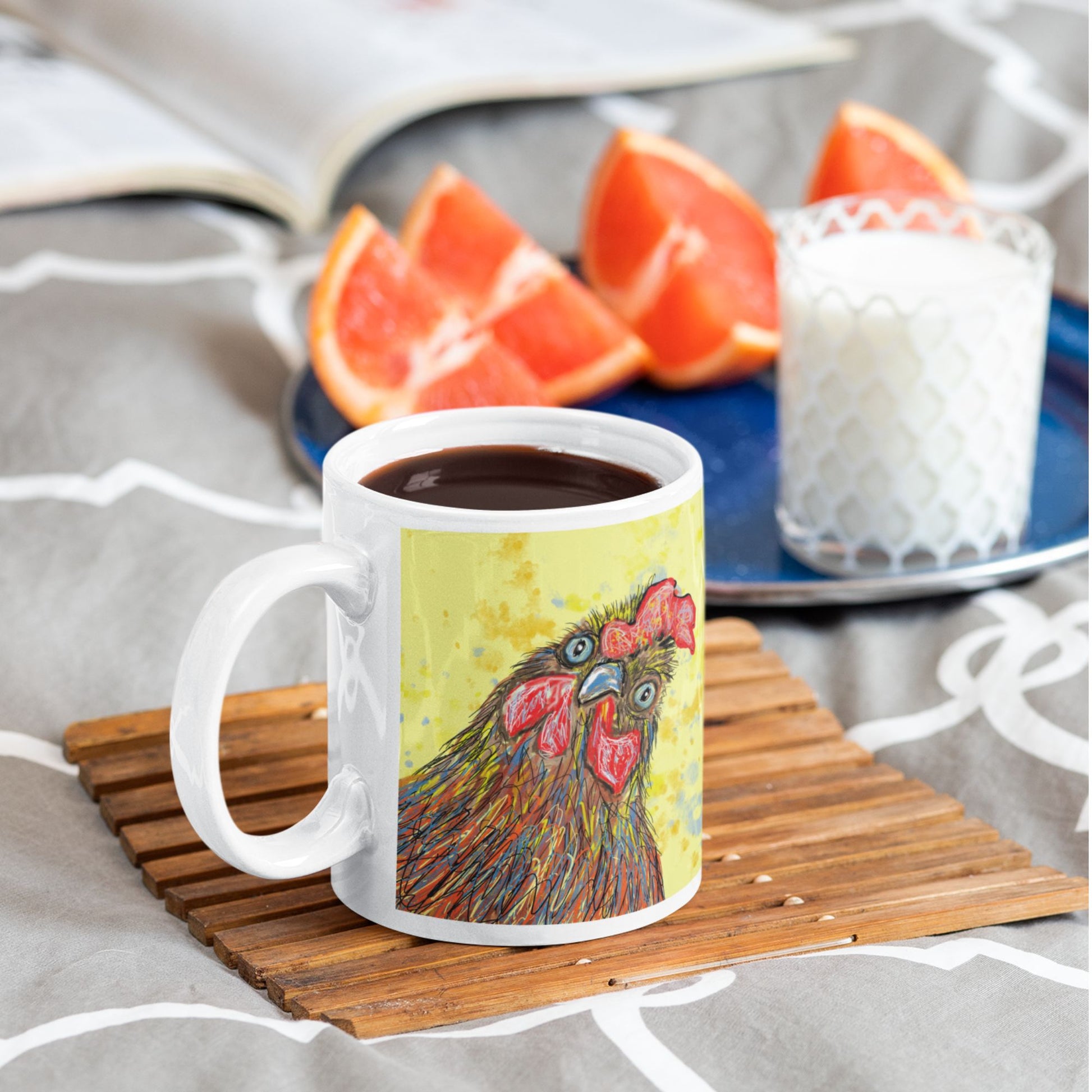 Mug with a rooster design on a wooden coaster, surrounded by on an orange with and a glass of milk.