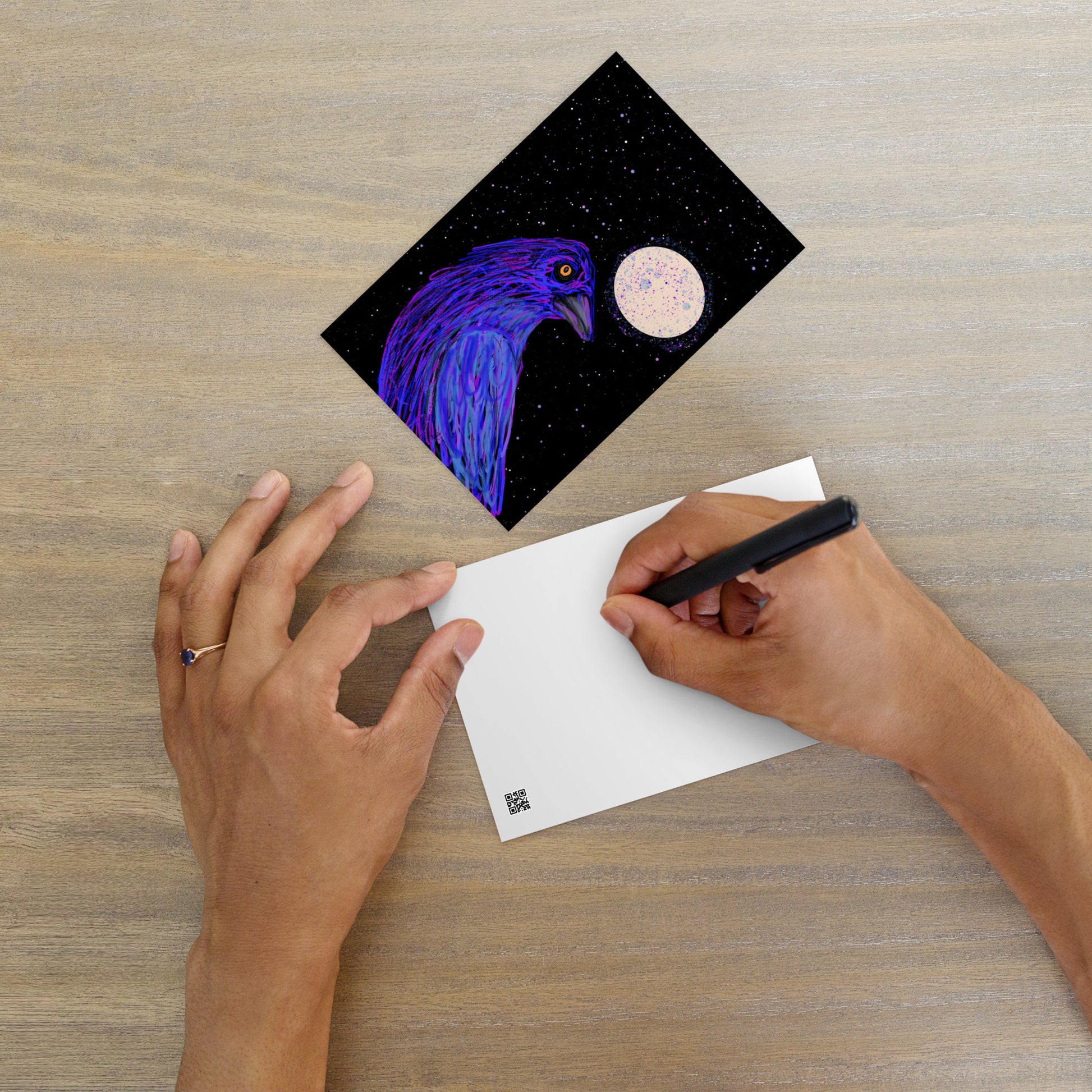 Crow with full moon and stars on a matte postcard. Crow symbolizes magic and mystery. Image shows someone writing on the back of the postcard, which is blank. 