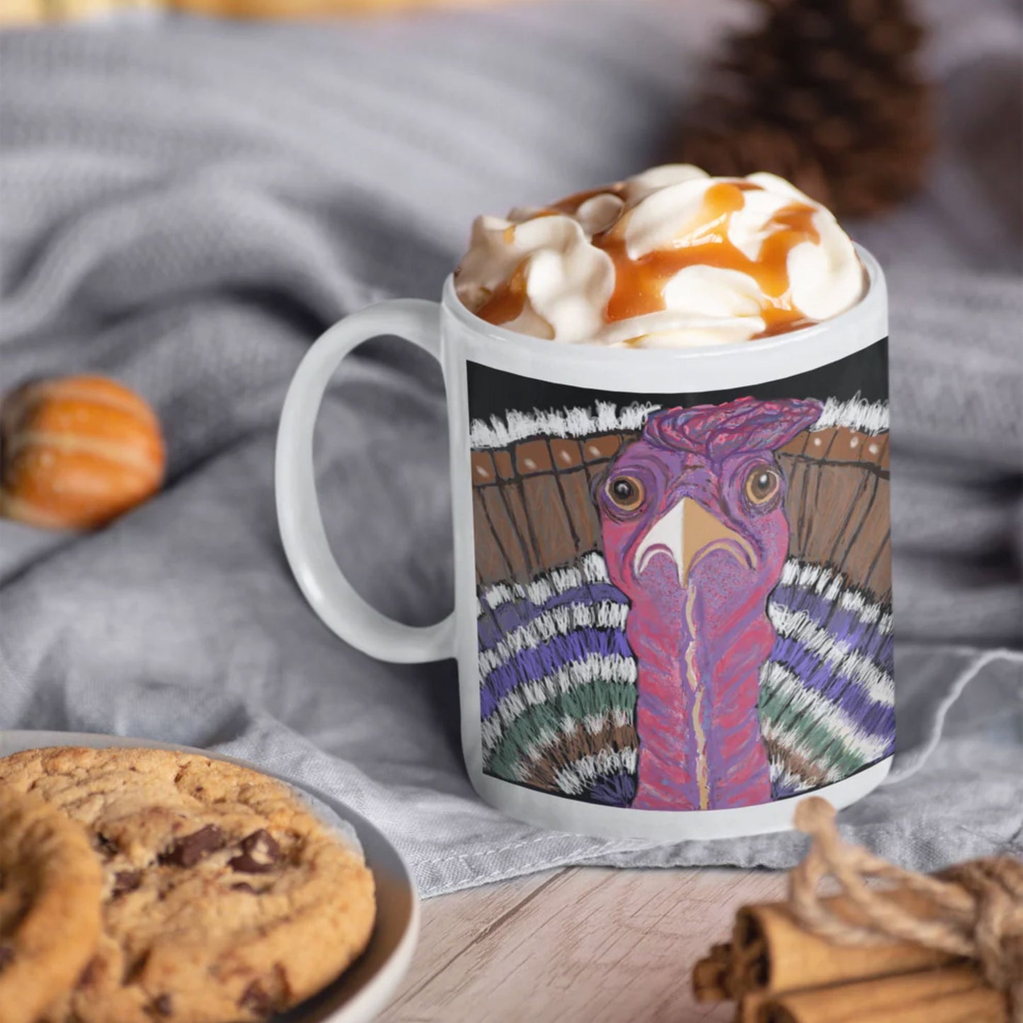 A ceramic turkey spirit mug with a colorful, original artists design filled with hot chocolate and whipped cream, placed on a wooden surface with cookies and cinnamon sticks.
