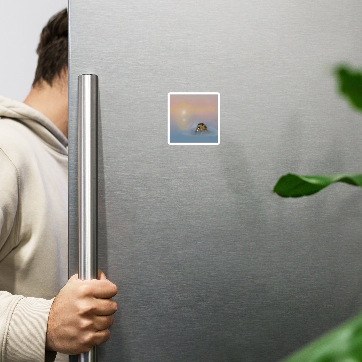 Person holding a silver refrigerator door open. There is a square magnet with an image of a walrus in a misty sea.
