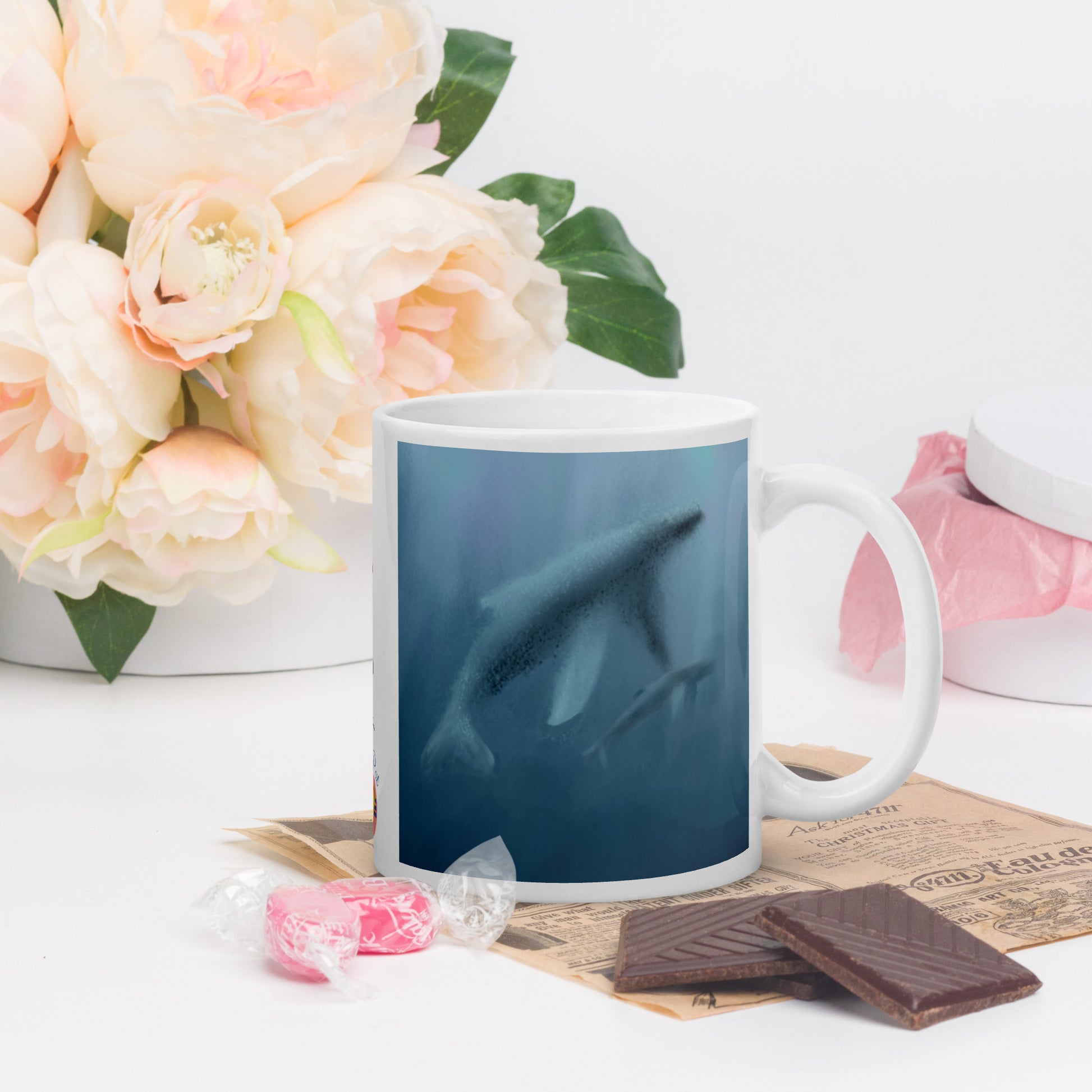Mug with a whale design on a table with flowers and chocolate. Misty blues showing mother and calf.