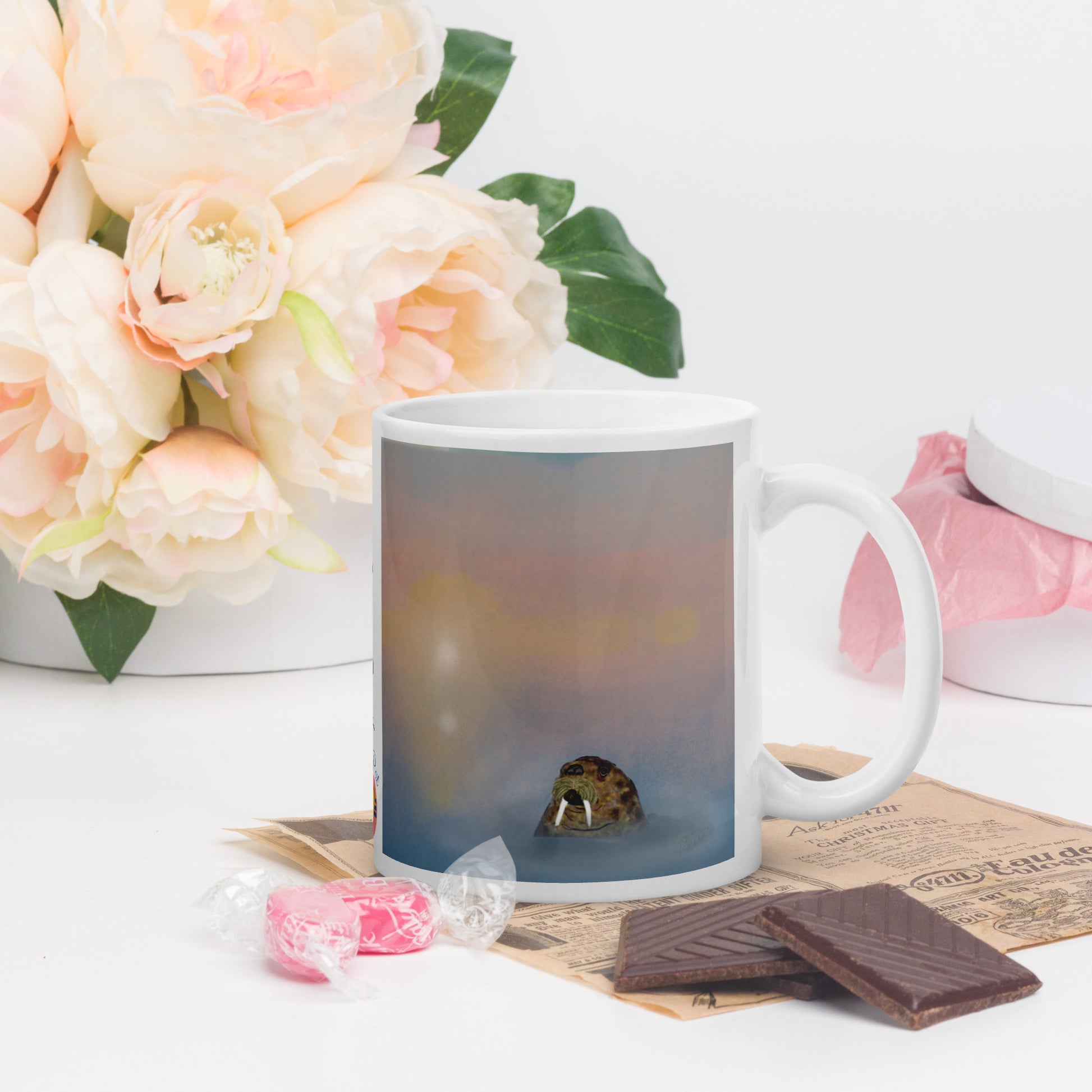 White mug with a printed design of a walrus in the water on a table with flowers and snacks