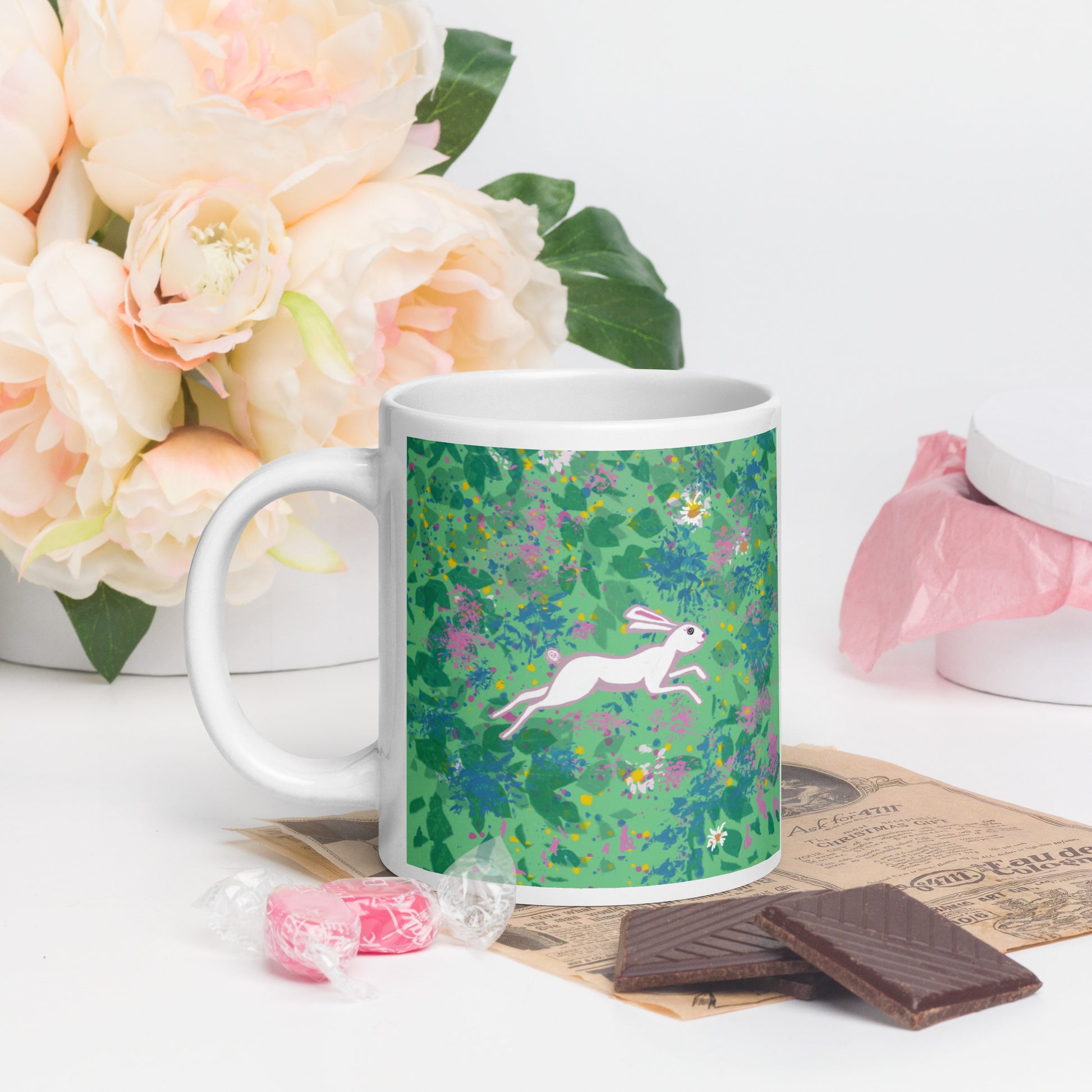 Mug with a floral design featuring a rabbit, placed on a table with flowers and chocolate.