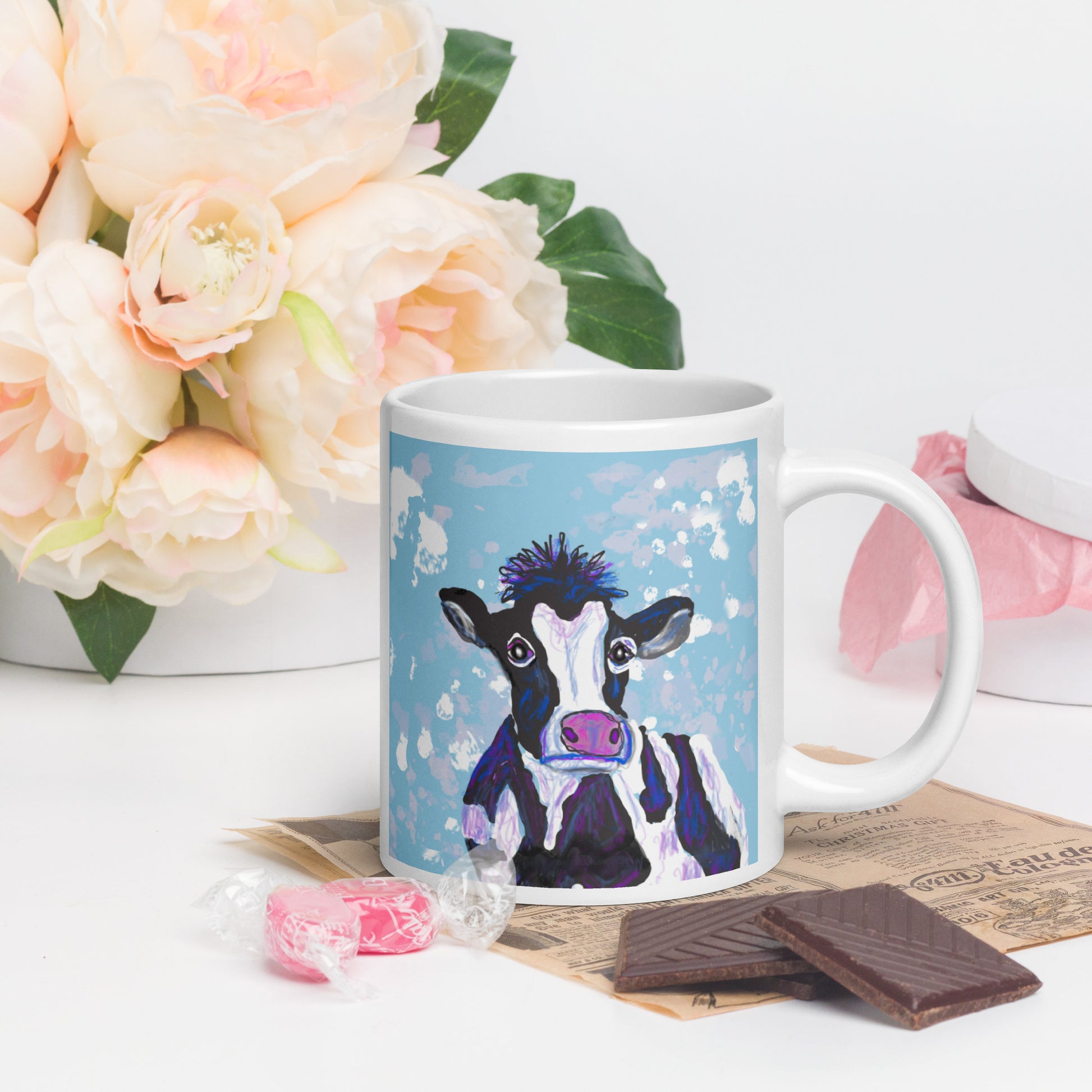Mug with a cow design on a table with flowers and chocolate. Cow symbolizes nurturing, abundance, and the gentle strength of Earth.
