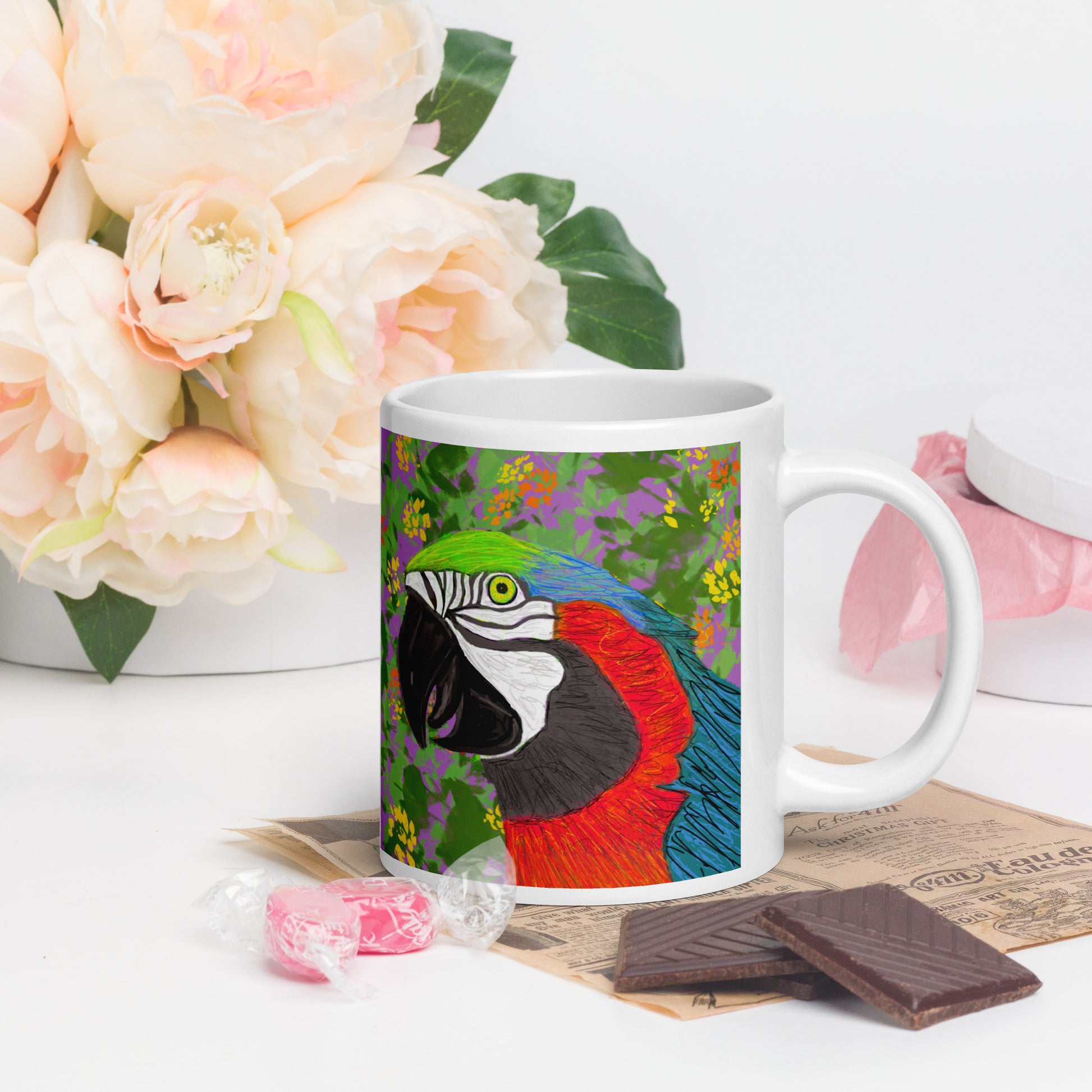 Mug with a colorful parrot design on a table with flowers and chocolates.