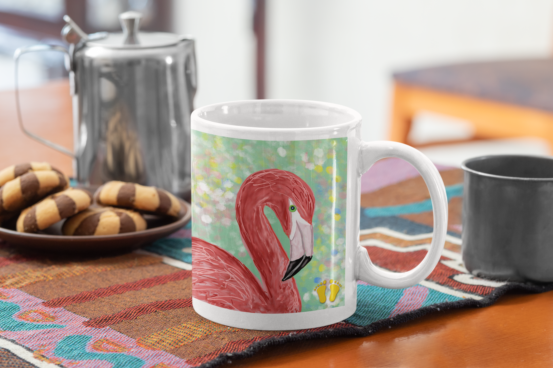 Premium ceramic coffee mug is a unique gift for bird lovers. Brightly colored flamingo is on a field of green with accents of white, pink, blue, and yellow. Shown here on a table with a tea pot and cookies. 