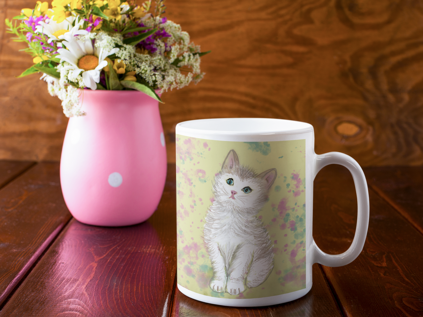 Premium ceramic coffee mug is a unique gift for cat lovers.  White kitten is on a field of yellow with accents of pink and green. Shown here on a table with a pink vase of mixed flowers.