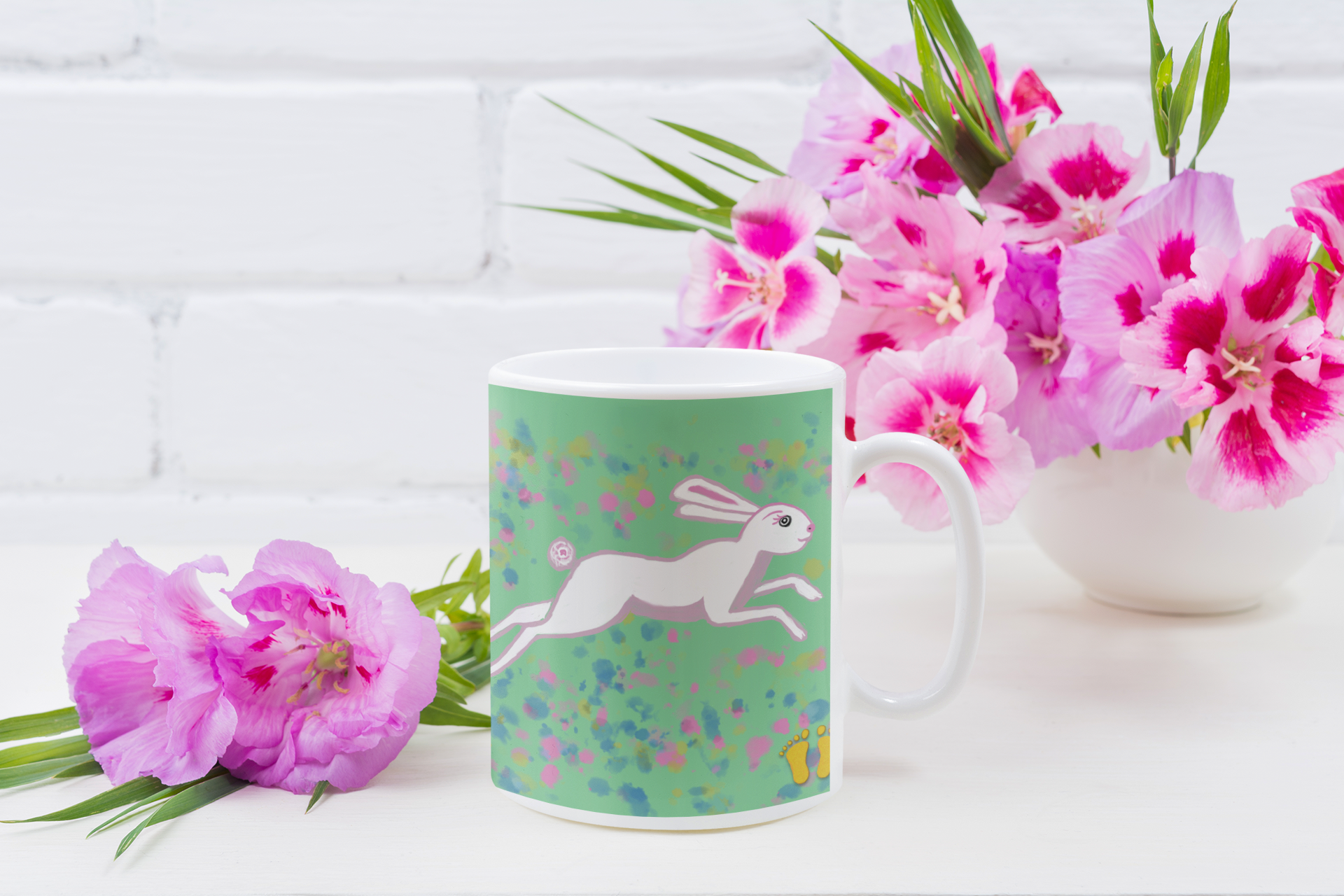 Unique gift for wild animal lovers. Cute March hare premium quality white ceramic coffee mug on a green, pink and blue field by PittaPat with the golden feet logo front view on white table with pink flowers.