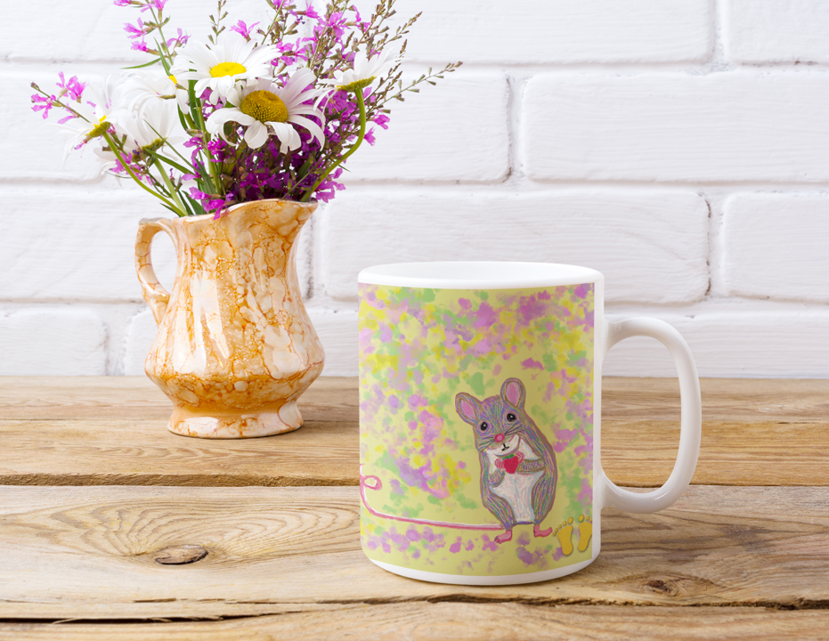 Premium ceramic coffee mug is a unique gift for animal lovers. Cute mouse holding strawberry premium quality white ceramic coffee mug on a yellow, pink and blue field by PittaPat. Front view on wood table with gold vase of flowers.