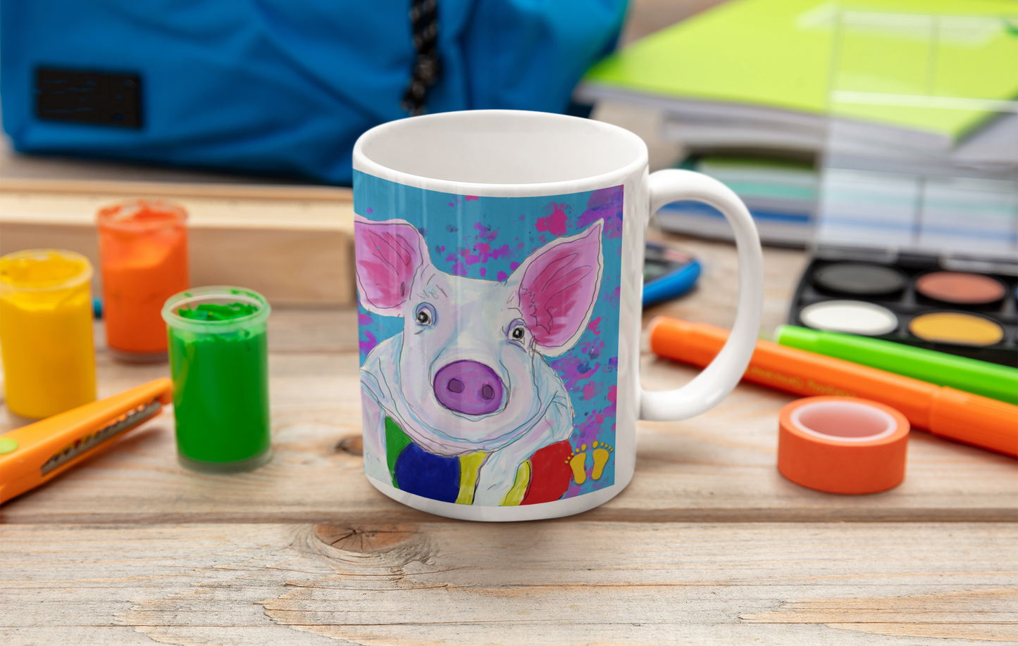 Colorful premium quality white ceramic coffee mug with a pig leaning on a ball on a blue and pink field by PittaPat with the golden feet logo sitting on a desk with art supplies