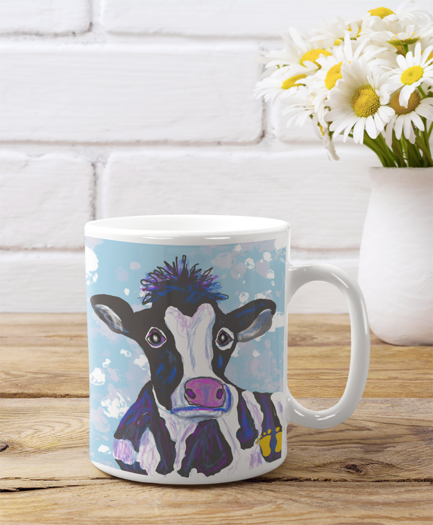 Premium ceramic coffee mug is a unique gift for animal lovers. Brightly colored cow is on a field of blue and yellow. Shown here on a table with a vase of daisies.