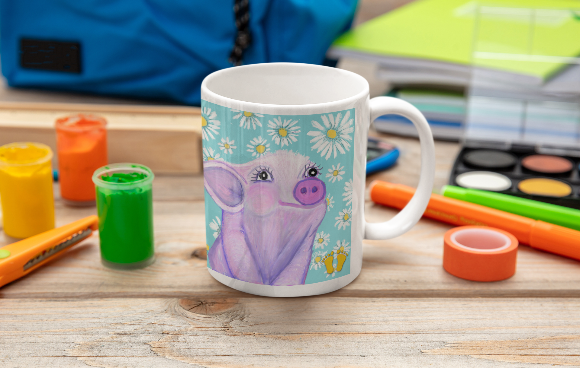 Premium ceramic coffee mug is a unique gift for animal lovers. Brightly colored piglet is on a field of blue with white and yellow daisies. Shown here on a table with artists’ paints and tools.